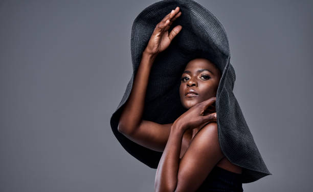 A woman poses confidently against a plain gray background, wearing a dramatic oversized black hat that frames her face and shoulders. Her expression is powerful and poised, with subtle, elegant makeup highlighting her features. The pose, combined with the striking hat, creates a bold and artistic fashion statement.