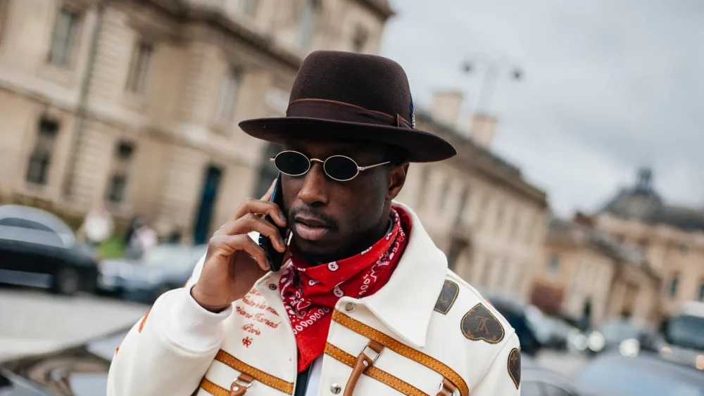 A man is captured outdoors in a stylish streetwear look. He wears a wide-brimmed brown hat, small round sunglasses, and a red bandana around his neck. His jacket is white with bold decorative patterns and leather details, giving a unique, fashionable edge. He is holding a phone to his ear, looking composed and confident, with a city backdrop of historic architecture behind him.