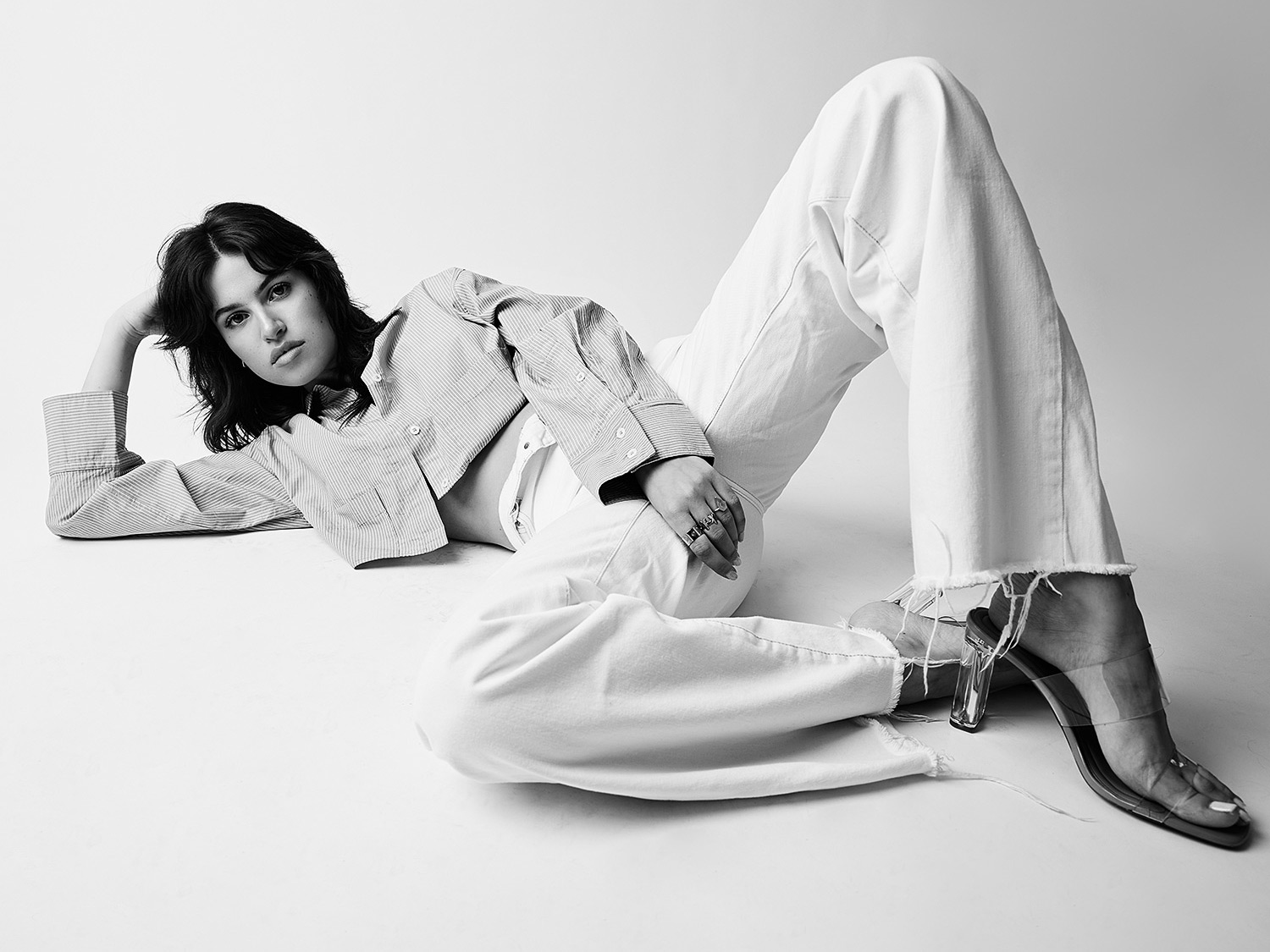 This black-and-white photo features a woman reclining in a confident, relaxed pose. She is dressed in wide-leg white pants with frayed hems, a cropped striped shirt, and clear high-heeled sandals. The minimal studio background emphasizes her modern, effortless style, while the monochrome tones highlight the sharp lines and textures of her outfit.