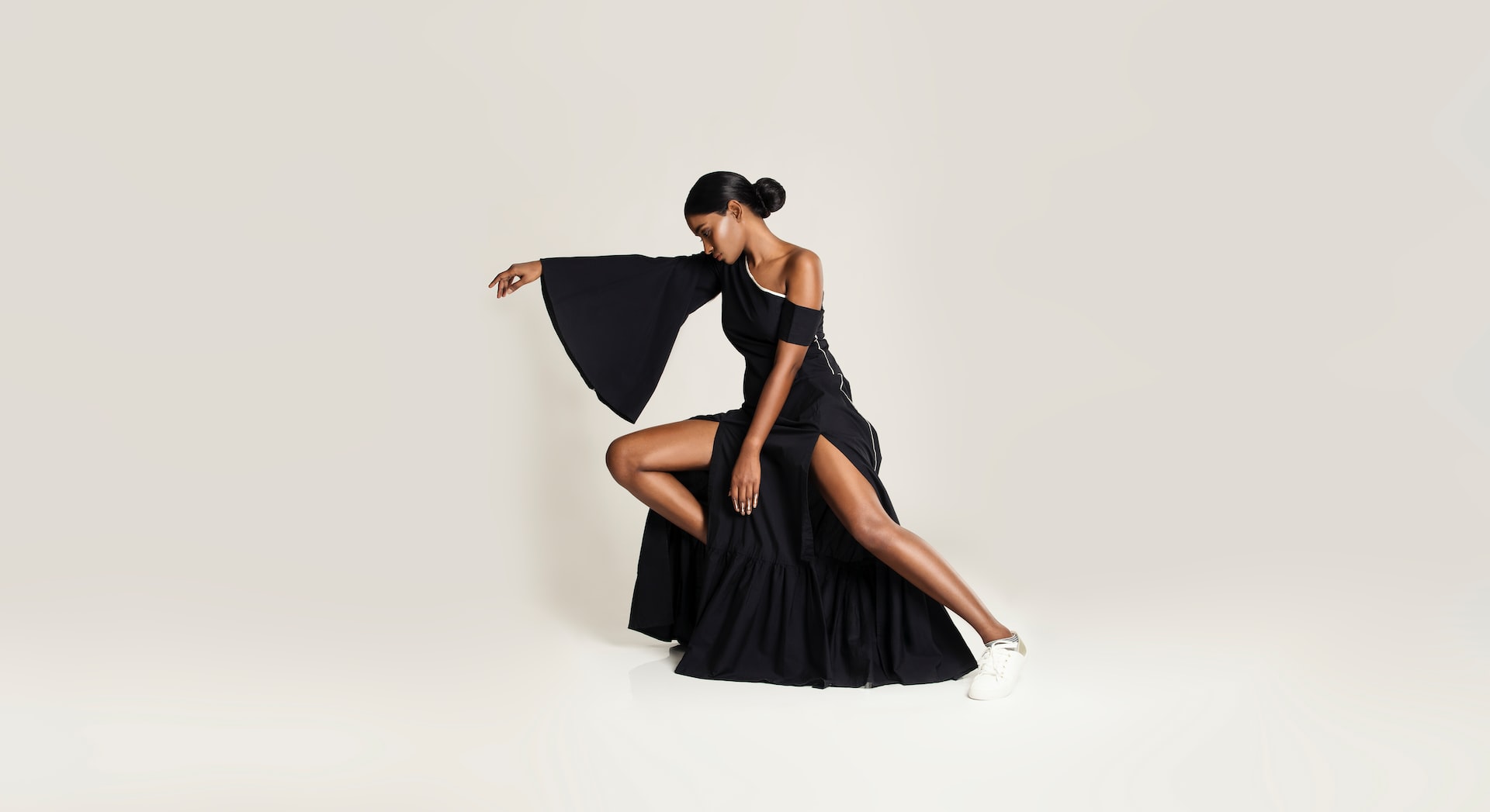 The photo features a woman in a striking black asymmetrical dress with dramatic flared sleeves and high slits. She is posed gracefully, extending one arm forward while resting on one bent knee, which emphasizes her long, elegant silhouette. The look contrasts the sophistication of the dress with the casual touch of white sneakers, creating a bold blend of high fashion and modern street style. The minimal beige background keeps the focus entirely on her and the dramatic styling.