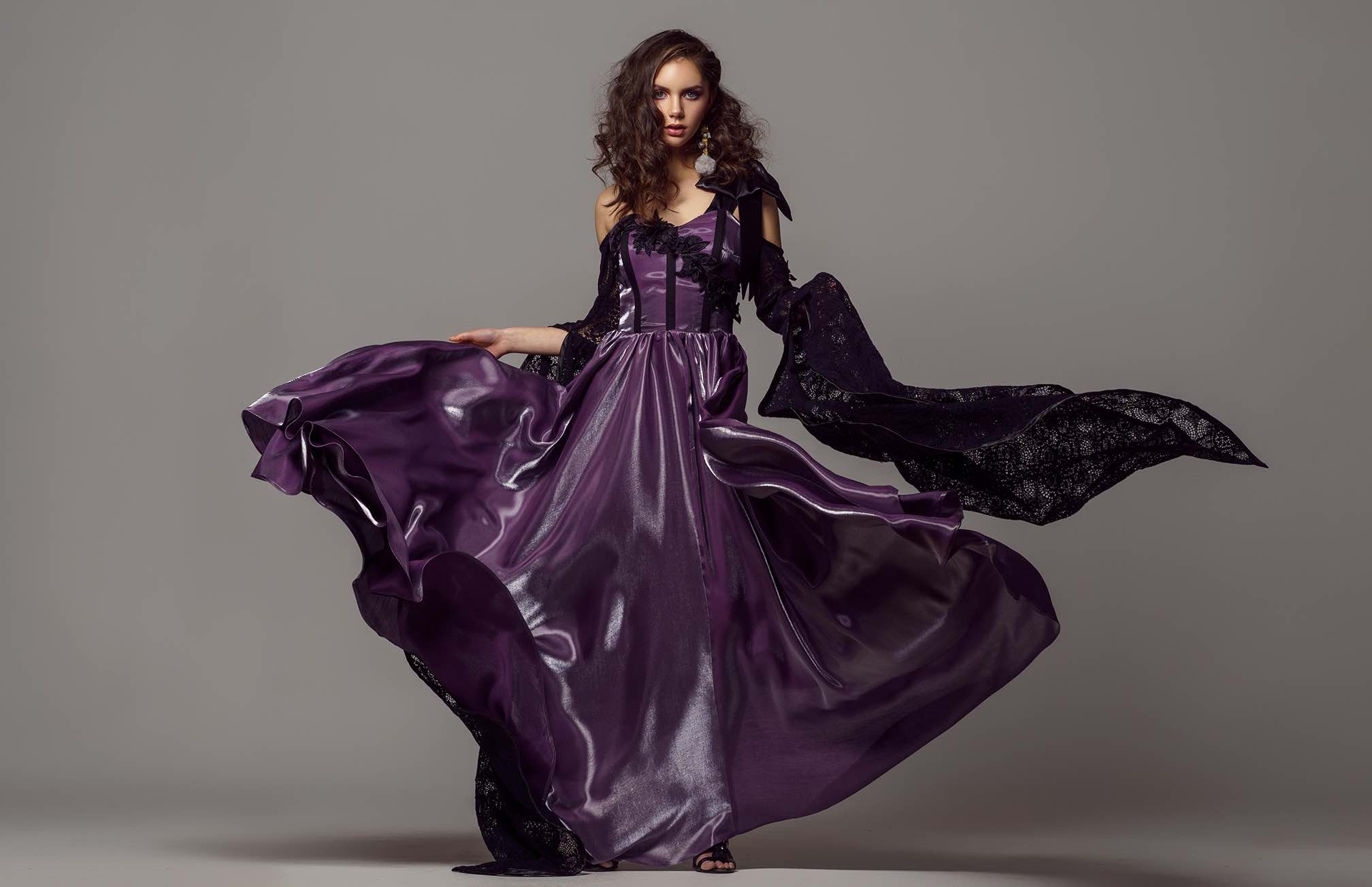 The photo shows a woman in a dramatic purple gown made of flowing, shiny fabric that spreads out beautifully around her as she moves. The dress has black lace details and an off-shoulder design, adding a touch of elegance and mystery. Her curly hair, statement earrings, and confident pose emphasize a bold and glamorous look. The neutral gray background highlights the rich color and movement of the dress.