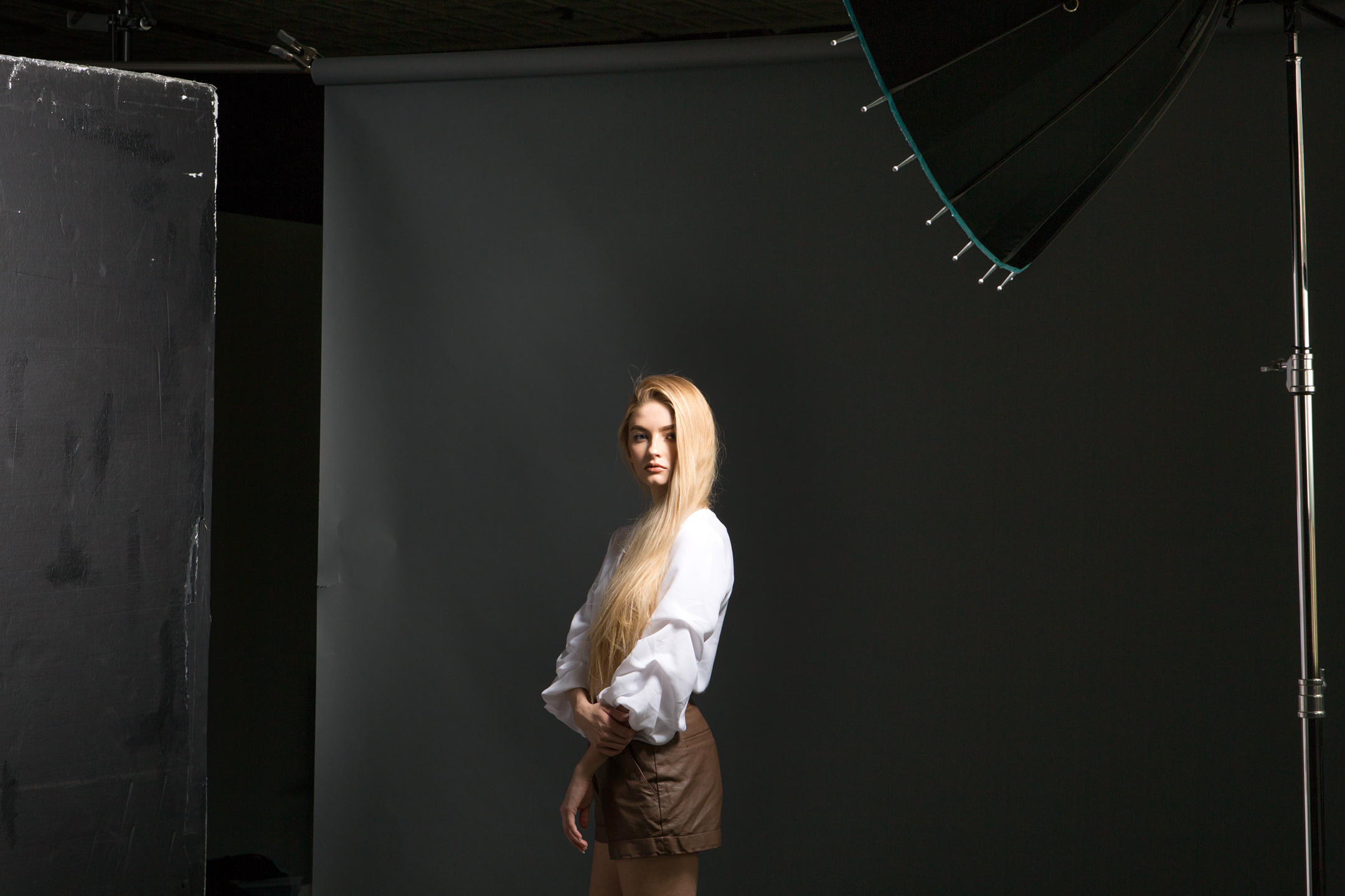 A young woman with long blonde hair poses in a studio setting. She is wearing a white blouse with puffed sleeves and brown high-waisted shorts. The background is dark, with professional studio lighting equipment partially visible, giving the photo a behind-the-scenes fashion shoot atmosphere.