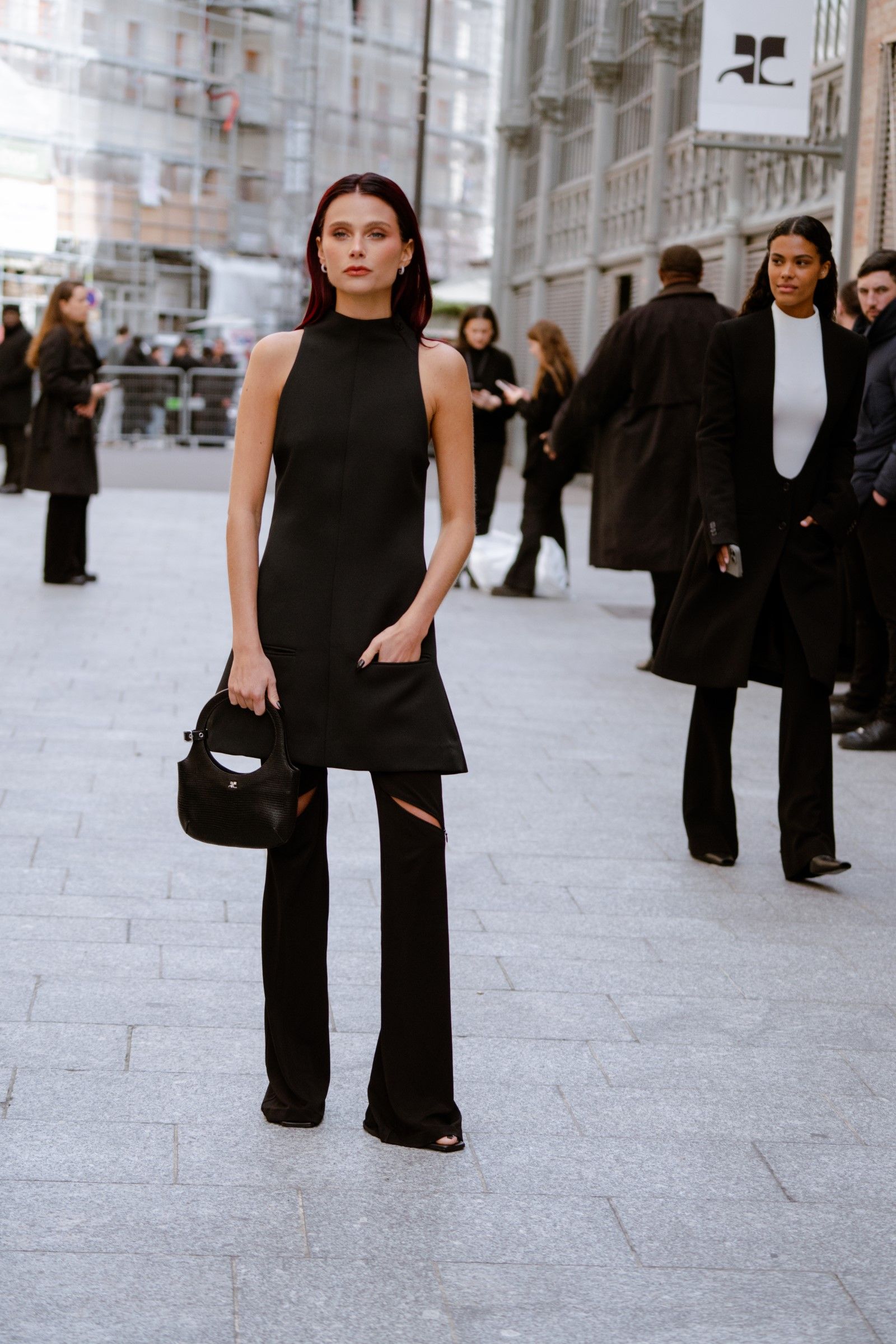 The photo shows a woman standing confidently on a city street, wearing a sleek all-black outfit. She pairs a sleeveless, high-neck tunic-style top with unique black pants featuring cut-out details above the knees. Her look is accessorized with a small black handbag and subtle earrings, while her straight dark hair adds to the sharp, modern aesthetic. The overall vibe is chic, minimalistic, and fashion-forward, perfectly suited for a stylish urban setting.