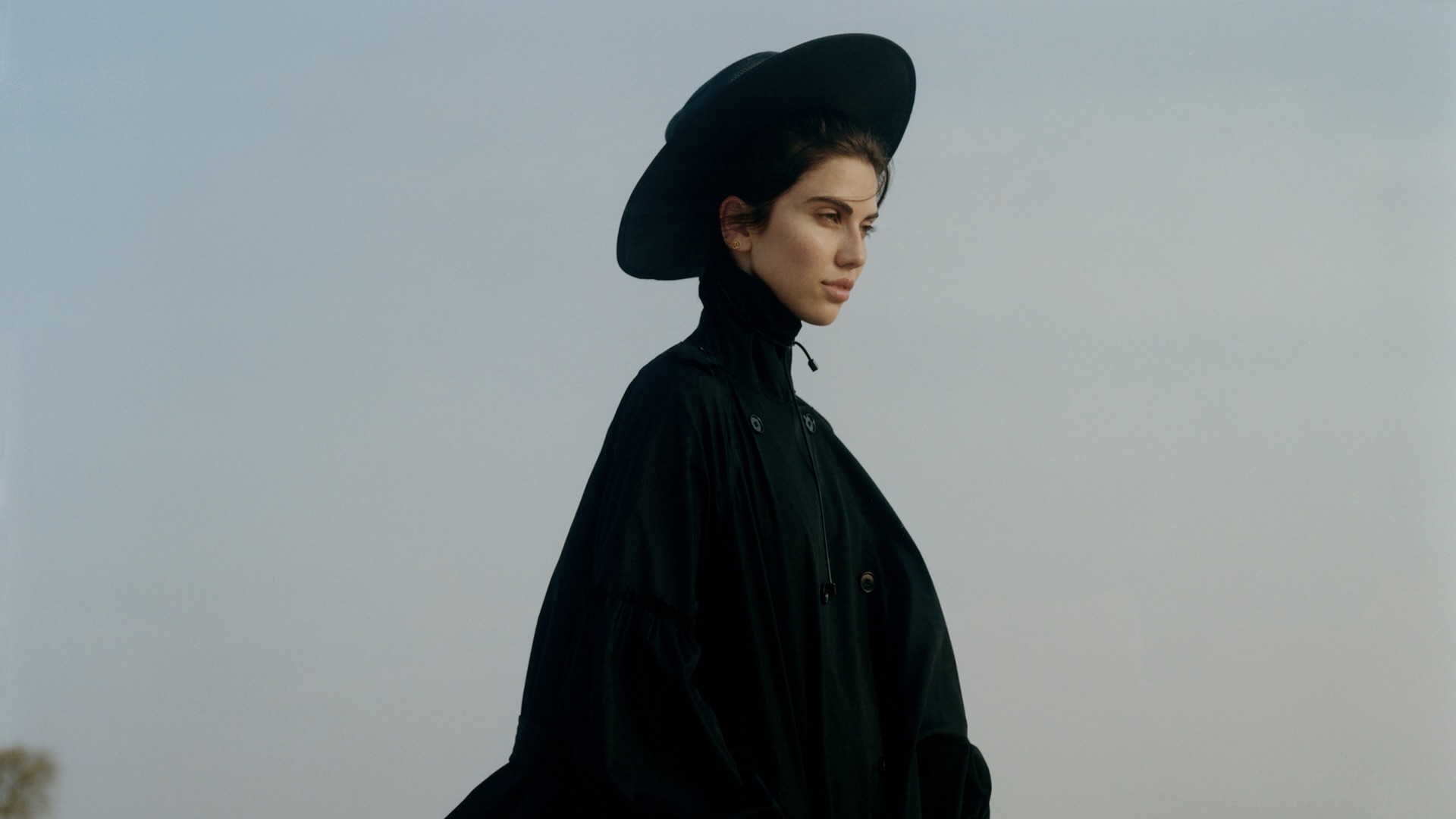 The photo shows a woman dressed in an all-black outfit, wearing a wide-brimmed hat and a high-collared coat. She stands against a soft, muted sky background, giving the image a calm, minimal, and slightly dramatic atmosphere. Her profile and composed expression add a sense of strength and elegance to the portrait.