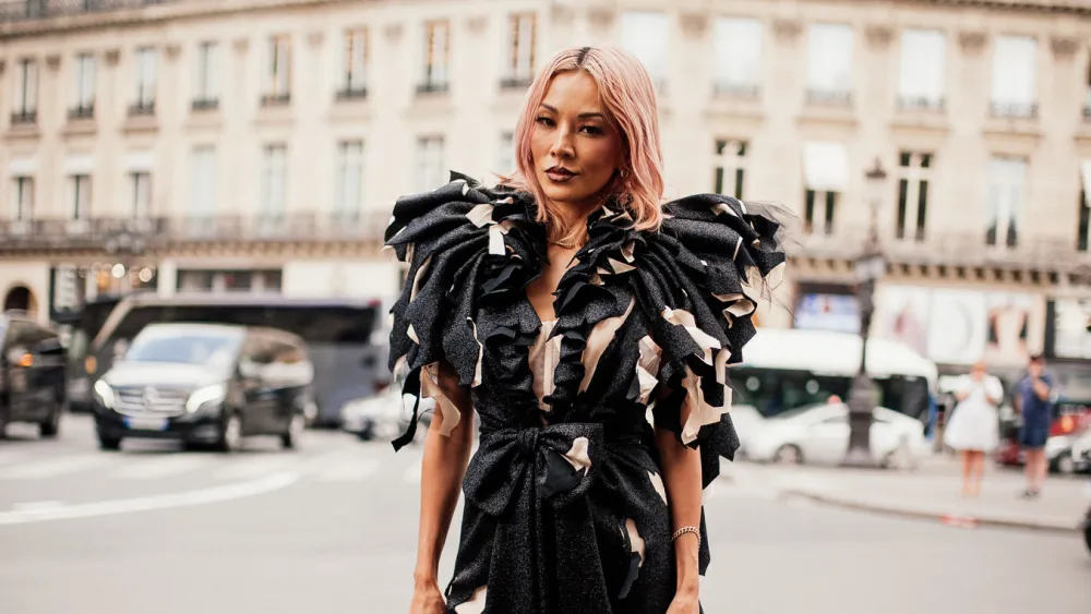 A woman stands confidently on a city street, dressed in a striking black outfit with bold ruffled and textured details. Her hair is styled in soft waves with a pinkish tone, and her makeup is polished and modern. The urban background with cars and classic architecture contrasts with her avant-garde fashion look, making her the clear focal point of the scene.